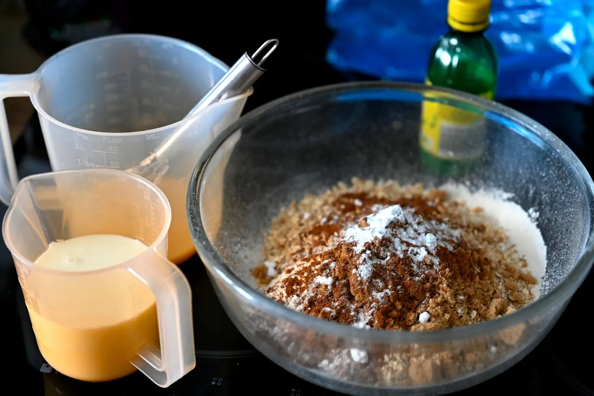 Glasschüssel mit Mehl, Zucker und Zimt, daneben Messbecher mit Flüssigkeit und eine Flasche, Zutaten zum Backen.