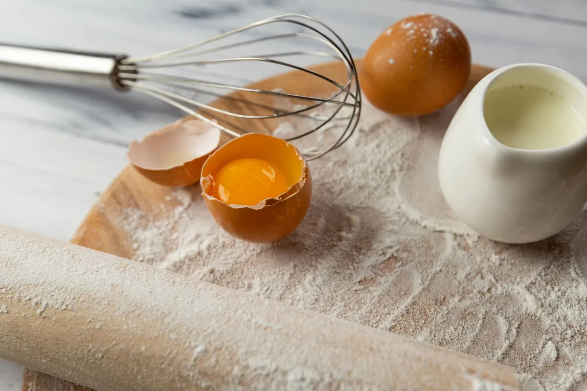 Aufgeschlagenes Ei, Schneebesen, Milchkännchen und Mehl auf einem Holzbrett mit Nudelholz