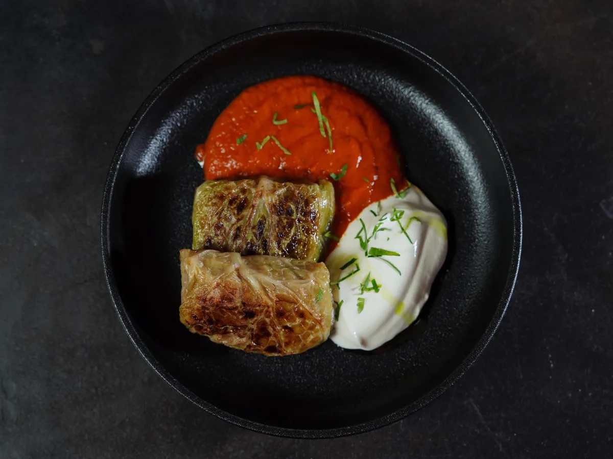 Zwei gebratene Kohlrouladen auf schwarzem Teller mit roter Tomatensauce und Joghurt- oder Sauerrahmklecks mit Kräutern