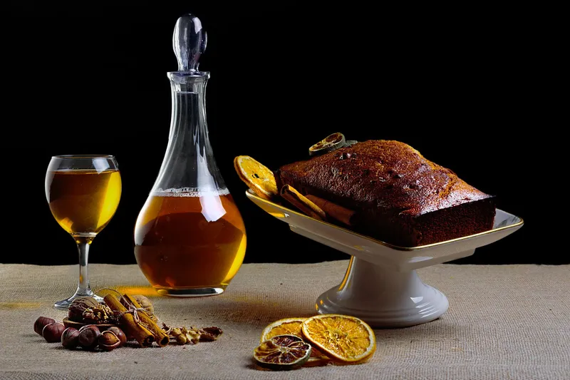Dunkler Kastenkuchen mit glänzender Oberfläche auf weißer Etagere, daneben Orangenscheiben, Zimtstangen.