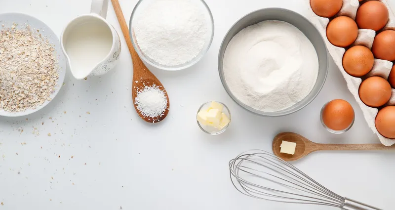 Backzutaten auf weißer Fläche: Eier im Karton, Mehl, Zucker, Butter, Milch, Haferflocken und ein Schneebesen